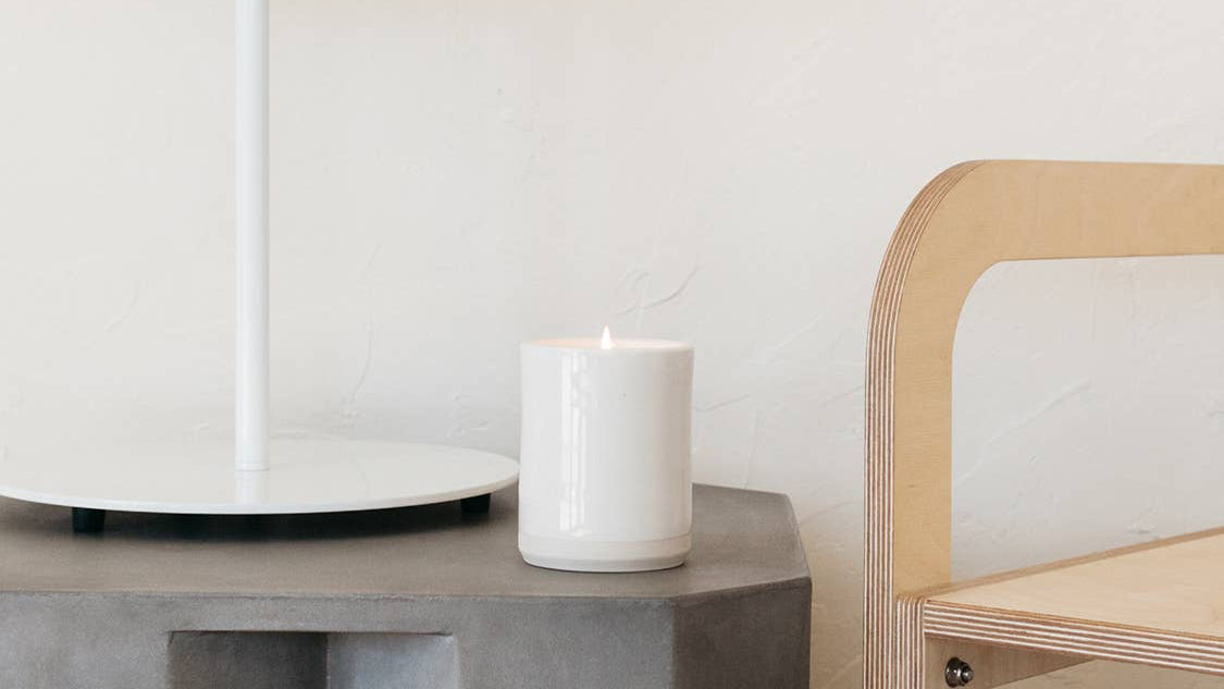 White candle on a concrete surface next to a wooden chair with a white wall background