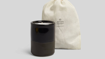 Black candle in a glass container with a beige drawstring bag on a light gray background