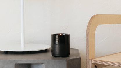 Black candle on a concrete surface next to a white lamp and wooden chair.